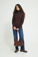 A woman modeling a dark brown Ichi Roll neck sweater, blue jeans, and brown boots, holding a brown suede slouchy shoulder bag against a plain light gray studio background.