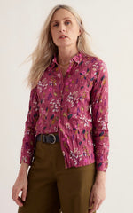 A woman modelling a Seasalt Larissa long-sleeve button-up shirt with a pink and purple floral print, tucked into olive green trousers with a black belt, against a plain light background.