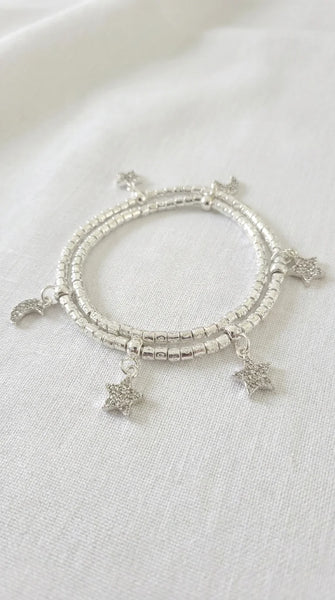 A wraparound silver beaded bracelet with dangling celestial charms, including stars and a crescent moon, displayed on a textured white fabric background.