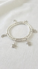 A wraparound silver beaded bracelet with dangling celestial charms, including stars and a crescent moon, displayed on a textured white fabric background.