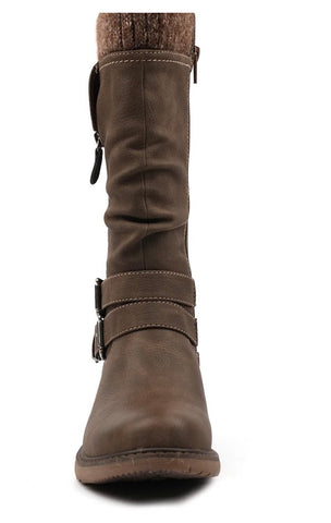 A person wearing Lunar Footwear taupe brown boots with buckle straps and side zippers, seated outdoors in snow beside a tree trunk.