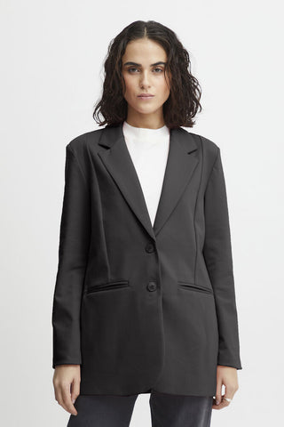 Close-up of a black blazer jacket with two dark buttons, a welt pocket, and a hand visible at the side, against a light neutral background.