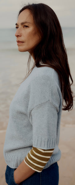 A woman modelling Seasalt light blue knit sweater with olive green and white striped elbow top, standing on a sandy beach with the ocean in the background.