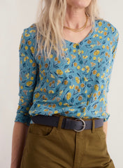 A woman wearing Seasalt blue 3/4 sleeve blouse with a yellow and green floral pattern, tucked into olive green pants with a dark belt.