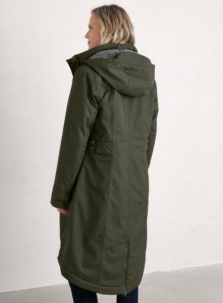 A woman wearing a mid-length olive green hooded jacket, shown from a side-back angle against a plain off-white wall.