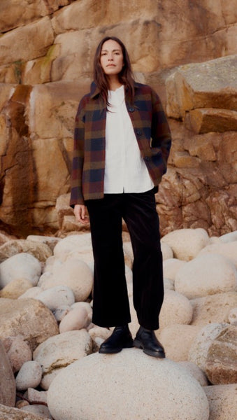 A woman modelling a Seasalt wool check coat, with a white blouse, black trousers and black boots. Stood behind a rock backdrop on a large stone. 
