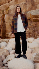 A woman modelling a Seasalt wool check coat, with a white blouse, black trousers and black boots. Stood behind a rock backdrop on a large stone. 