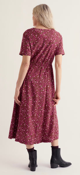 A woman shown from the back wearing Seasalt short sleeve midi dress with a dense small floral print in burgundy, pink, and yellow, paired with black ankle boots, against a plain light beige studio background.
