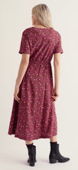 A woman shown from the back wearing Seasalt short sleeve midi dress with a dense small floral print in burgundy, pink, and yellow, paired with black ankle boots, against a plain light beige studio background.