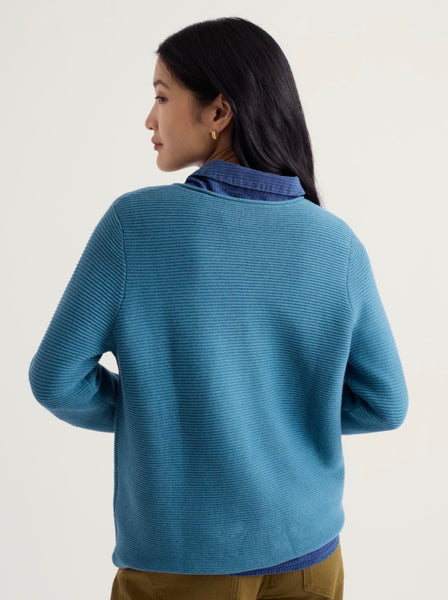 A woman shown from the back wearing a Seasalt blue ribbed knit sweater over a blue collared shirt, paired with olive brown pants, against a plain light background.