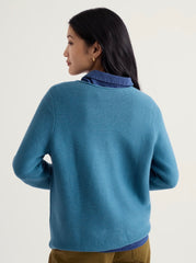 A woman shown from the back wearing a Seasalt blue ribbed knit sweater over a blue collared shirt, paired with olive brown pants, against a plain light background.