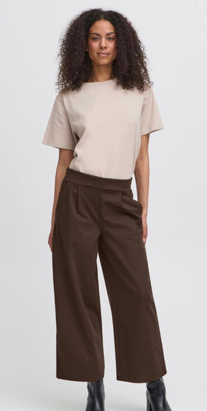 A model with curly dark hair is wearing a beige short-sleeve top and Ichi dark brown wide-leg cropped trousers, paired with black boots, against a light grey studio background.