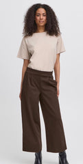 A model with curly dark hair is wearing a beige short-sleeve top and Ichi dark brown wide-leg cropped trousers, paired with black boots, against a light grey studio background.