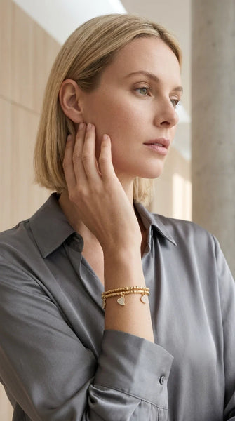 Women wearing a grey blouse modelling an Envy gold wraparound bracelet with heart and circle diamanté charms. Against a studio background.