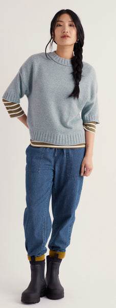 A woman modelling Seasalt light blue knit sweater with striped sleeves and hem, paired with blue jeans and black boots, against a plain light background.