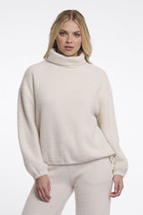 A woman wearing Rino & Pelle cream coloured turtleneck sweater with matching sweatpants against a plain white studio background.