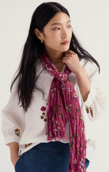 A woman wearing a Seasalt white embroidered blouse and blue jeans, styled with a pleated magenta scarf featuring a small floral print.
