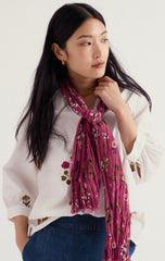 A woman wearing a Seasalt white embroidered blouse and blue jeans, styled with a pleated magenta scarf featuring a small floral print.