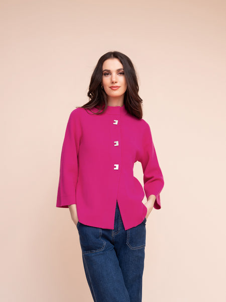 A woman modeling a bright magenta long-sleeve blouse with decorative white geometric buttons down the front, paired with dark blue jeans, against a solid pale peach background.