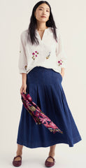 A woman modeling a white long sleeve Seasalt blouse with colourful floral embroidery on the chest and sleeves, paired with a dark blue pleated midi skirt and holding a pink floral scarf, against a plain light background.