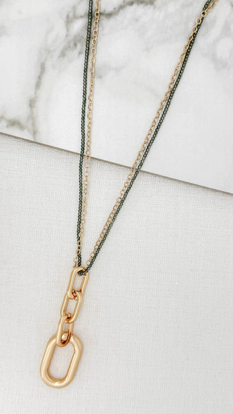 Envy gold-toned chain necklace with a large open-link pendant, layered over a thin dark green chain, displayed on a white textured surface with a marble accent.