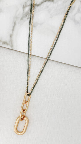 Envy gold-toned chain necklace with a large open-link pendant, layered over a thin dark green chain, displayed on a white textured surface with a marble accent.