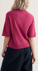 A woman shown from the back wearing Seasalt short sleeve knit sweater in a rich magenta pink colour, paired with dark navy trousers, against a plain light grey background.