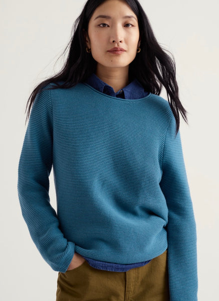 A woman wearing Seasalt blue ribbed knit crewneck sweater layered over a dark blue collared shirt, paired with brown trousers, against a plain light background.