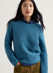 A woman wearing Seasalt blue ribbed knit crewneck sweater layered over a dark blue collared shirt, paired with brown trousers, against a plain light background.