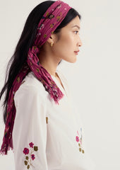 A woman in profile wearing a Seasalt magenta floral headscarf tied under her chin and a white embroidered blouse, photographed against a clean off-white background.