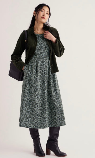 A woman models a dark green velvet jacket over a green and navy floral print midi dress, paired with black knee-high boots and a black shoulder bag, against a plain off-white studio background.