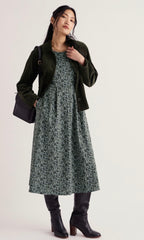 A woman models a dark green velvet jacket over a green and navy floral print midi dress, paired with black knee-high boots and a black shoulder bag, against a plain off-white studio background.