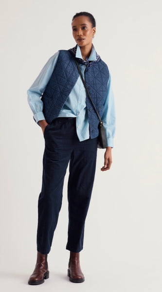 A model stands against a light grey studio background wearing a light blue button down shirt, a navy quilted vest, Seasalt  dark navy trousers, and brown leather boots. A black shoulder bag is slung across the body.