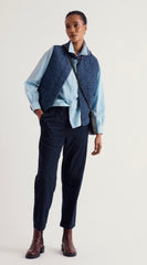 A model stands against a light grey studio background wearing a light blue button down shirt, a navy quilted vest, Seasalt  dark navy trousers, and brown leather boots. A black shoulder bag is slung across the body.