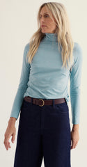 A woman wearing a Seasalt light blue long sleeve roll neck top with dark blue jeans, and a brown leather belt with a gold buckle, photographed against a plain off-white studio background. 