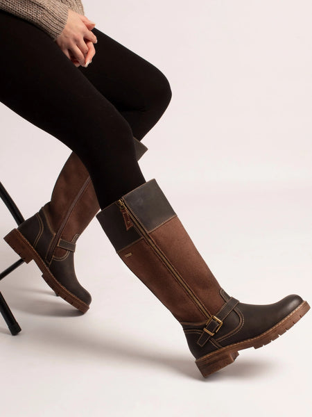 A person seated on a stool wearing tall brown boots with dark panels and buckle details, paired with black tights, against a plain light background.