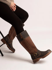 A person seated on a stool wearing tall brown boots with dark panels and buckle details, paired with black tights, against a plain light background.