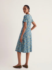 A woman modelling Seasalt short sleeve midi length dress with a blue and yellow floral pattern, shown from a side back angle against a plain light background.