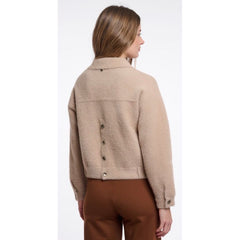 A woman shown from the back wearing a cropped beige  jacket with a button-down back and brown pants, against a plain white background.