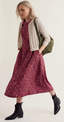 A woman modelling Seasalt long burgundy floral midi dress with a cream cable-knit cardigan, olive green shoulder bag, and black ankle boots against a plain light beige studio background.