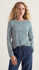 Model is wearing Seasalt Top in a ditsy print. Long sleeve fitted top paired with a pair of blue trousers with models hand in pocket in studio background.