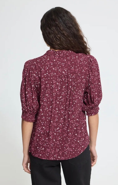 A woman viewed from the back wearing a burgundy short-sleeve shirt with a small white floral print and black pants, against a plain light grey studio background.