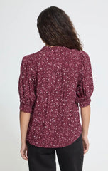 A woman viewed from the back wearing a burgundy short-sleeve shirt with a small white floral print and black pants, against a plain light grey studio background.