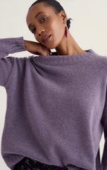 A woman wearing a Seasalt loose, oversized knit sweater in a muted dusty lavender-purple colour, paired with dark patterned bottoms and pink earrings, against a plain off-white background.