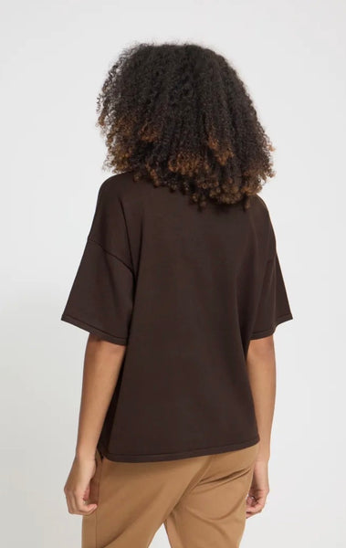 A person shown from the back wearing an oversized dark brown short-sleeve Ichi top and tan pants against a plain light gray studio background.