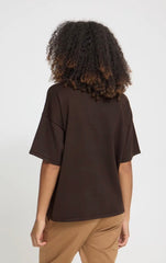 A person shown from the back wearing an oversized dark brown short-sleeve Ichi top and tan pants against a plain light gray studio background.