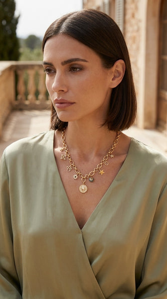 A woman with short dark brown hair wearing Envy Jewellery gold star and coin charm necklace over a sage green wrap top, set against a warm stone balcony background.