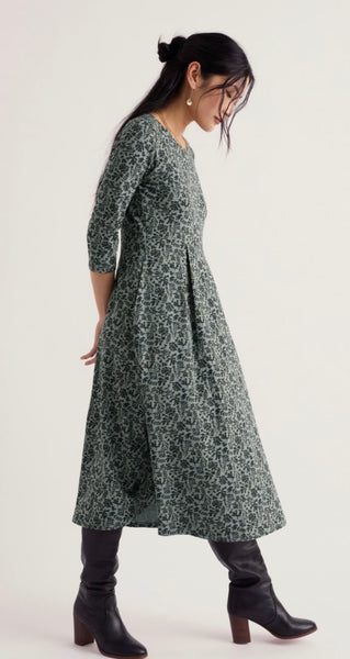 A woman wearing a Seasalt dark green and navy floral print midi dress with three-quarter sleeves, paired with black leather knee-high boots, photographed against a plain off-white studio background.