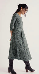 A woman wearing a Seasalt dark green and navy floral print midi dress with three-quarter sleeves, paired with black leather knee-high boots, photographed against a plain off-white studio background.