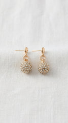 Envy Jewellery a pair of gold-toned dangle earrings with round, crystal-encrusted balls, displayed on a textured white fabric background.